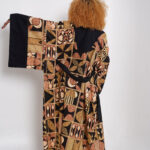 Woman wearing a long Afrincan kimono with bold tribal patterns in earthy tones, standing against a white background with one arm extended to show the wide sleeve.