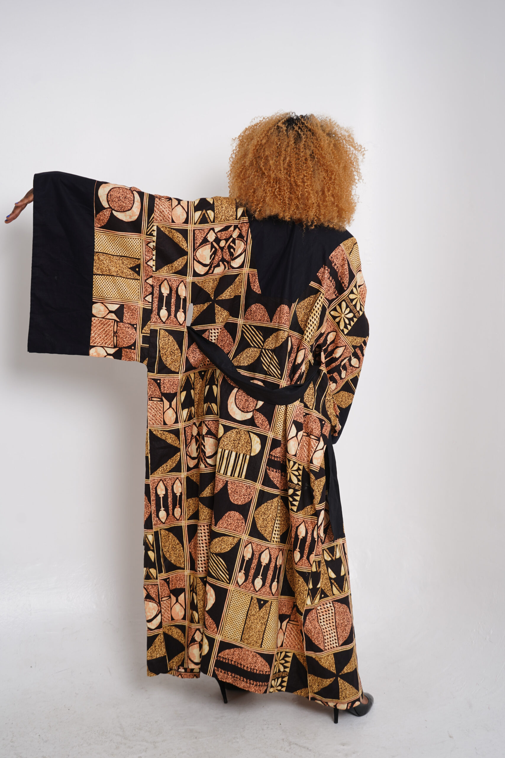 Woman wearing a long Afrincan kimono with bold tribal patterns in earthy tones, standing against a white background with one arm extended to show the wide sleeve.
